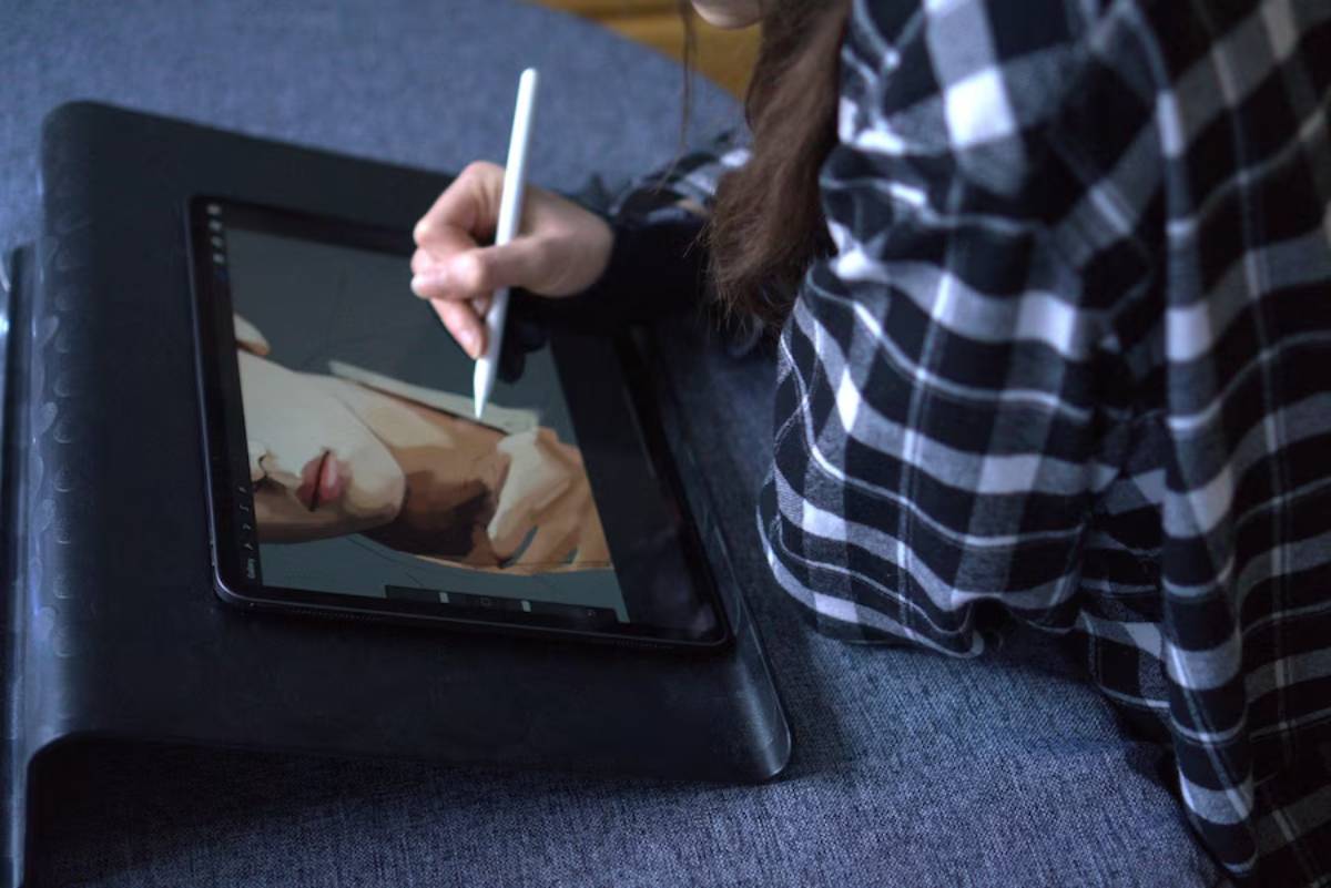 A person in a plaid shirt sketches a portrait on a tablet using a stylus, with focus on their hand and the digital artwork.