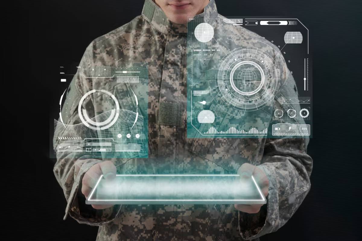 A soldier in camouflage holds a futuristic tablet, displaying holographic data and interface panels in a dark setting.