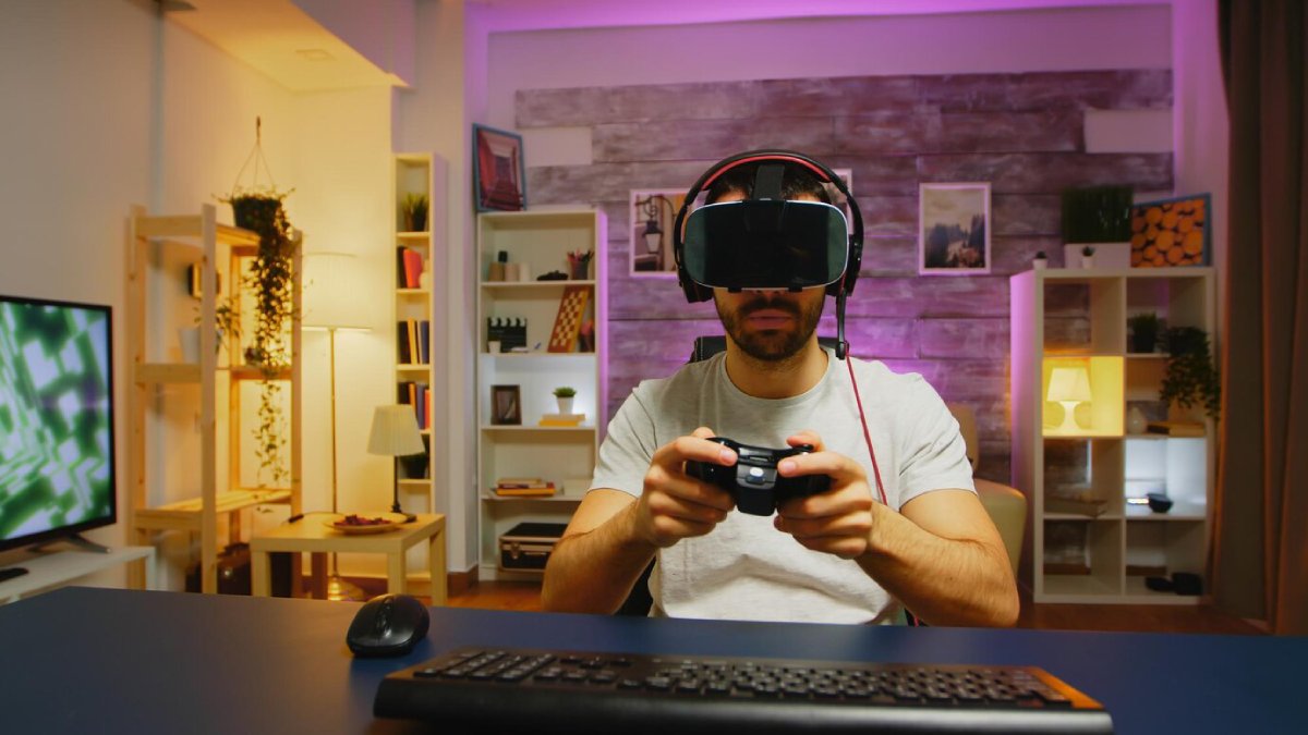 A gamer holds a game controller, seated at a desk with a computer mouse and keyboard, in a cozy, modern room with ambient lighting.