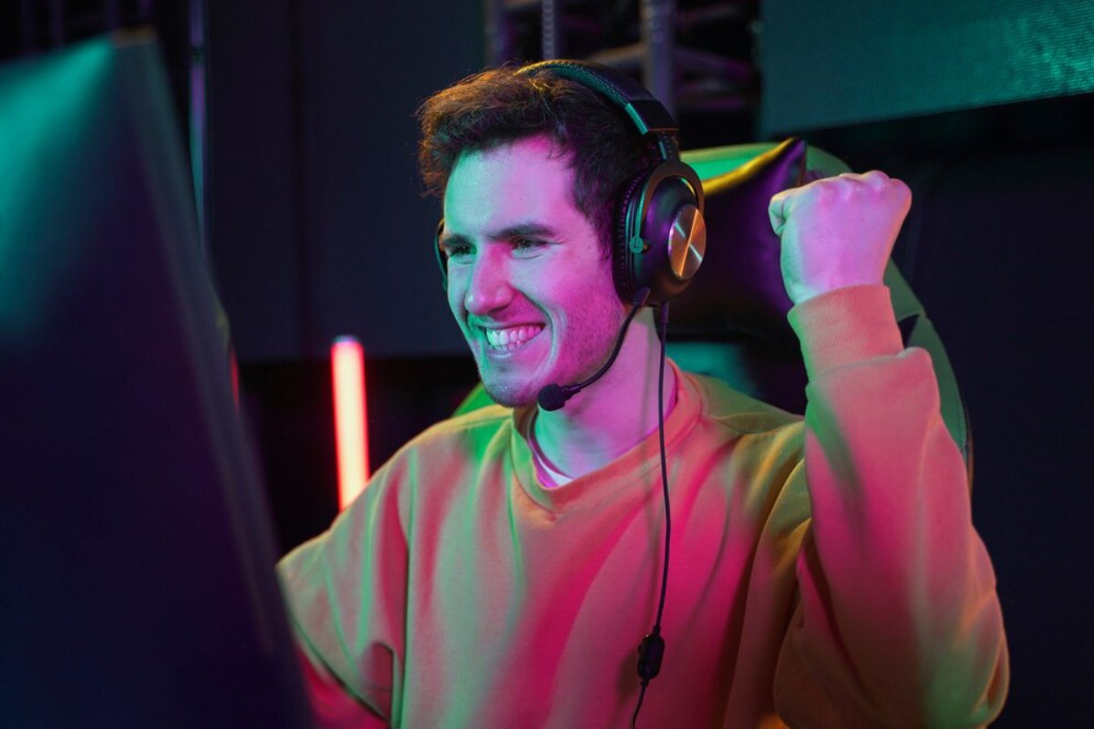 A gamer in a colorful setting, wearing headphones, celebrates with a raised fist while playing video games.