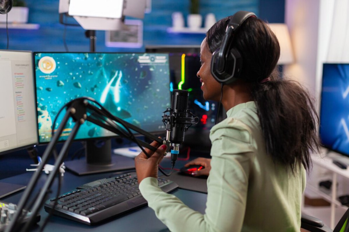 A person wearing headphones sits at a desk with two monitors, speaking into a microphone, engaged in gaming or streaming.
