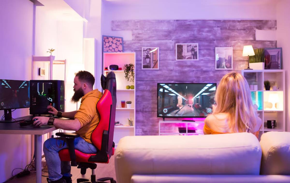 A cozy gaming setup with two people playing video games, one at a desk with dual monitors and the other on a couch, in a warmly lit room.