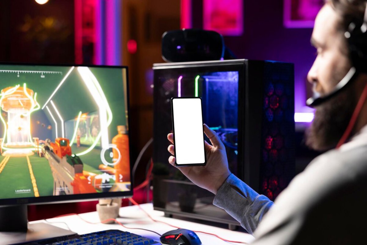 A person holds a smartphone displaying a blank screen, with a colorful gaming setup and a computer monitor showing an action-packed game.