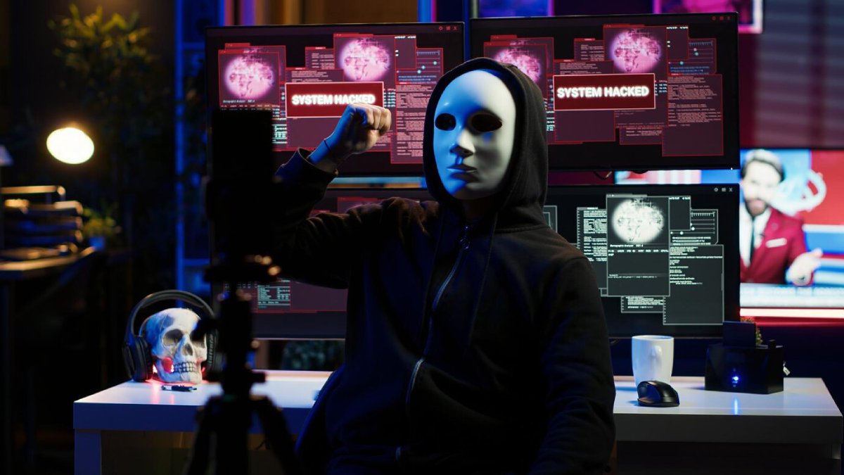 A person in a hoodie sits at a desk with multiple monitors displaying a SYSTEM HACKED message, alongside a skull and coffee mug.
