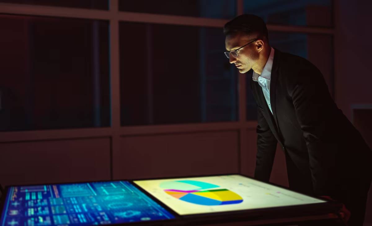 A person in a suit leans over a large digital display showing graphs and charts in a dimly lit office setting.