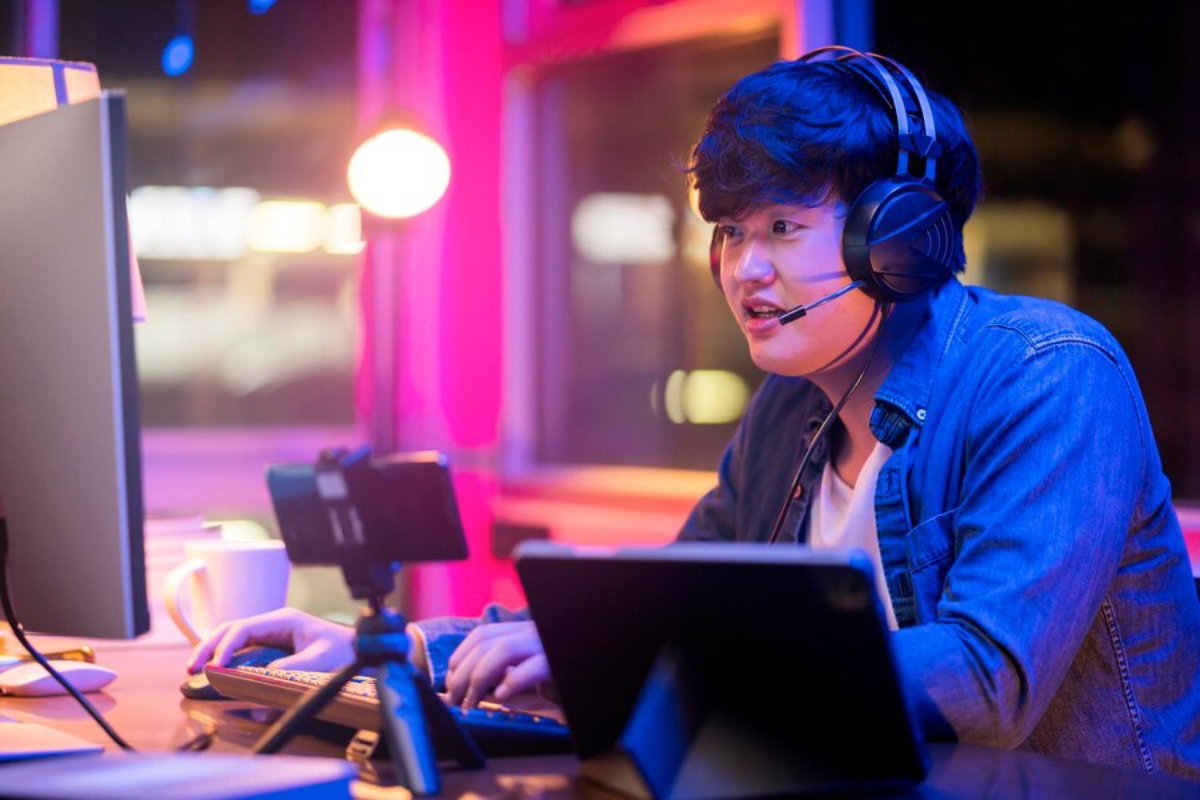 A person wearing headphones sits at a desk with a computer, a tablet, and a coffee mug, surrounded by colorful ambient lighting.