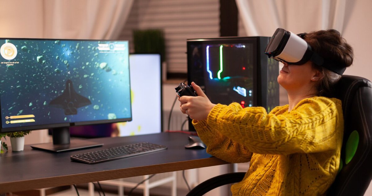 A person wearing a VR headset plays a space exploration game at a desk with dual monitors and a gaming PC, wearing a yellow sweater.