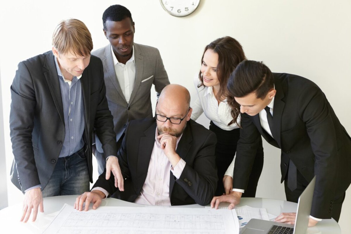 A diverse group of five professionals huddles around a table, examining blueprints and discussing plans. A laptop is visible nearby.