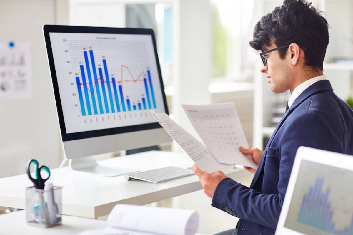 A person in a suit reviews documents while focused on a computer screen displaying colorful graphs and data analysis charts.