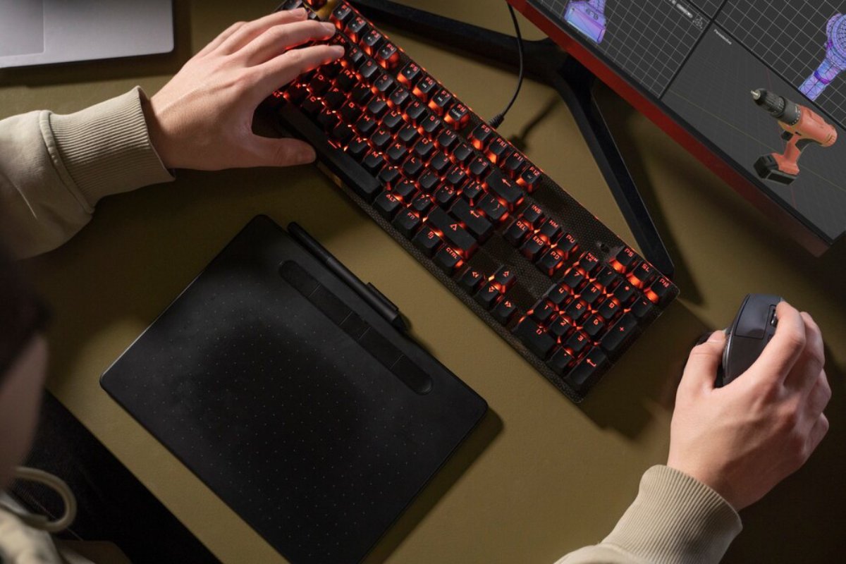 An overhead view of a person's hand on a gaming mouse, with a keyboard emitting red light and a drawing tablet nearby.