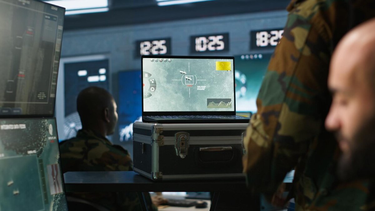A military operations room with a laptop display showing a targeting interface, flanked by monitors and personnel in camouflage.