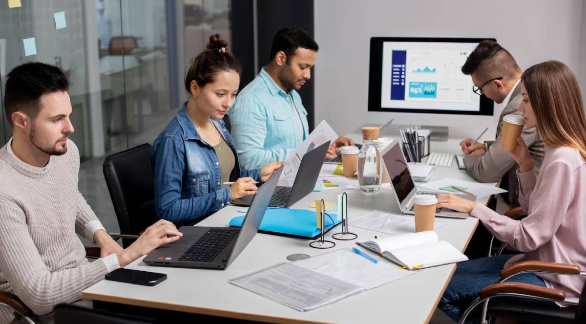 A diverse group engaged in a collaborative meeting, using laptops and discussing materials around a modern conference table.