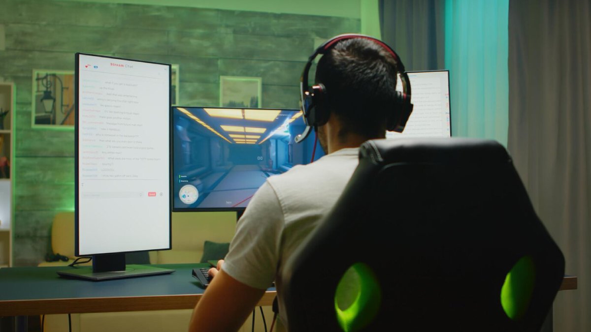 A gamer in a headset plays on dual monitors, one displaying a game scene and the other showing a live chat interface.