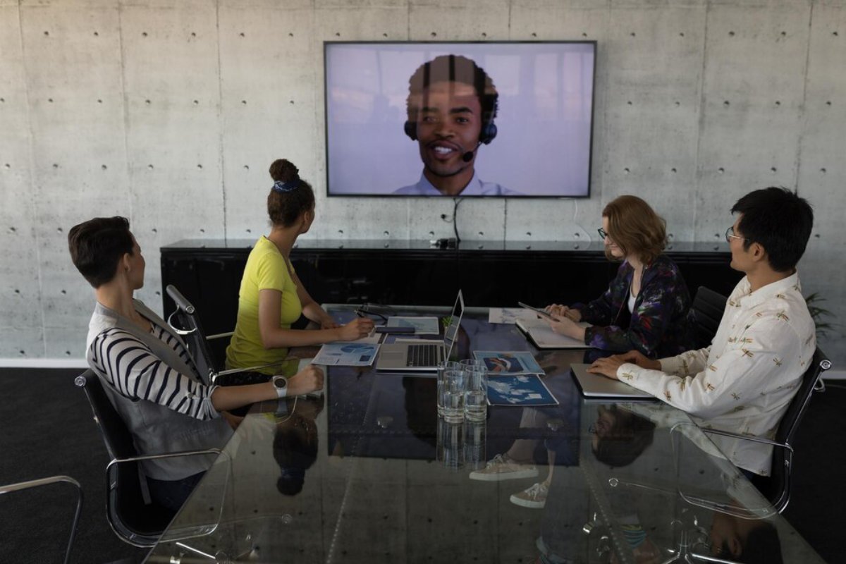A group of four people sit around a glass table in a modern conference room, engaged in a video call displayed on a wall-mounted screen.