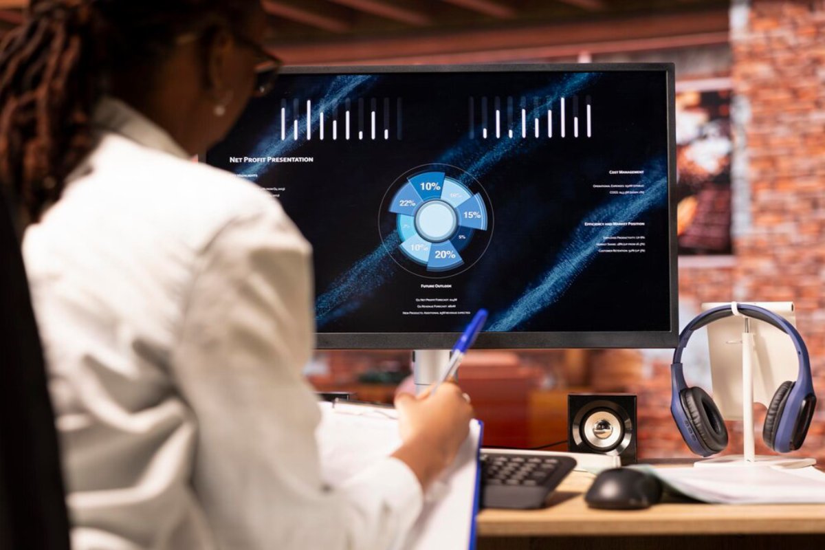A person with glasses reviews a net profit presentation on a computer screen, surrounded by headphones and a speaker on a desk.