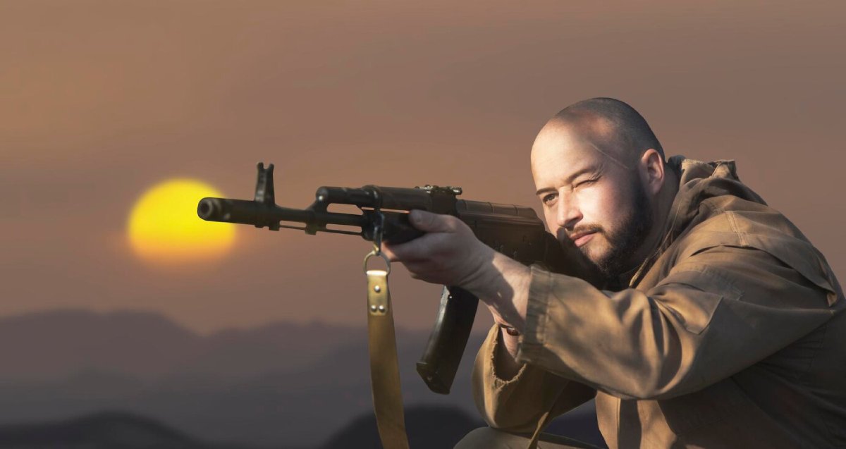 A person in military attire aims an assault rifle toward the horizon, with a vibrant sunset casting warm colors in the background.