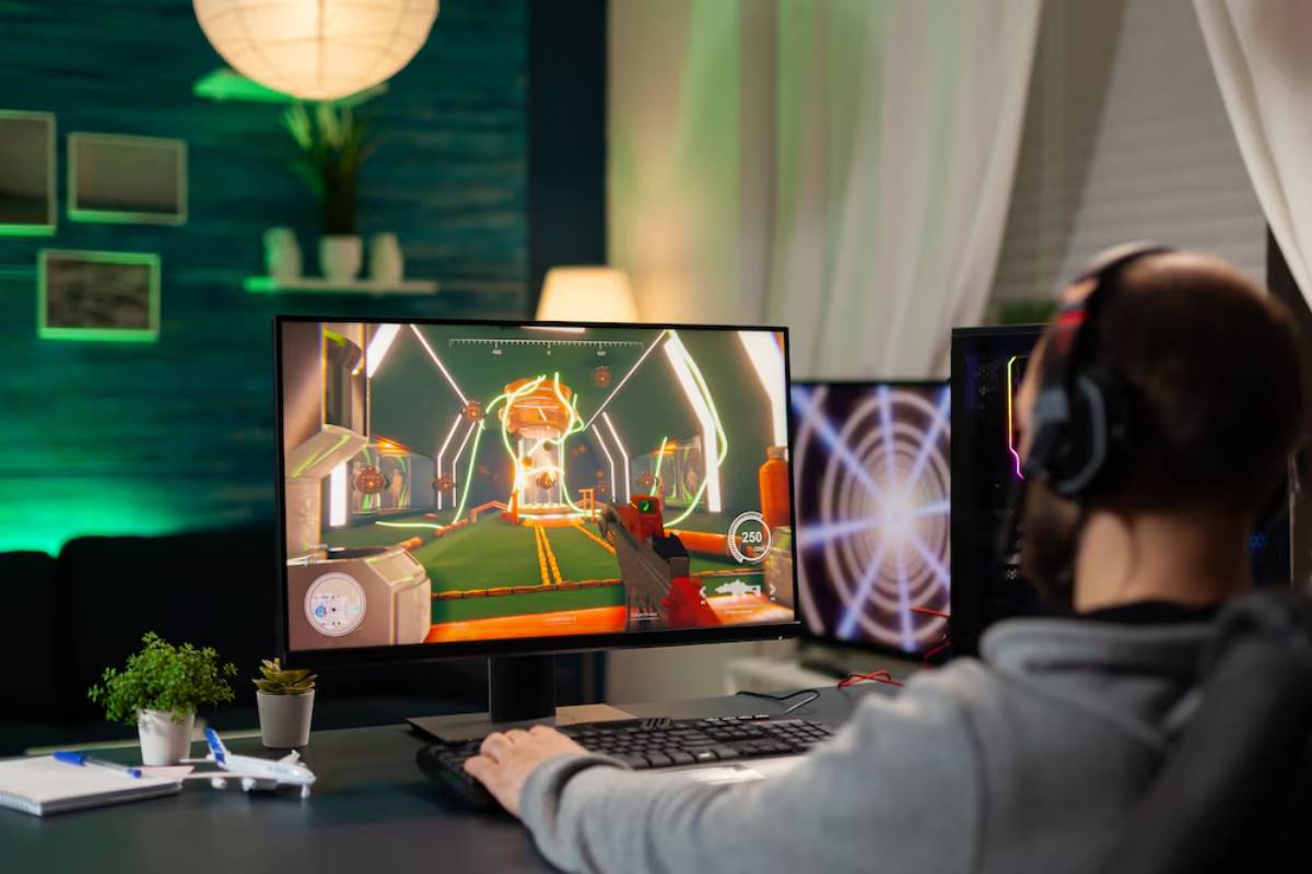 A gamer in a gray hoodie plays a video game on a monitor, wearing headphones in a cozy, dimly lit room with green accents.