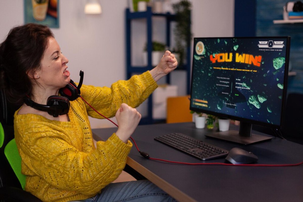 A person in a yellow sweater celebrating a gaming victory, sitting at a desk with a monitor displaying YOU WIN! and gaming equipment.