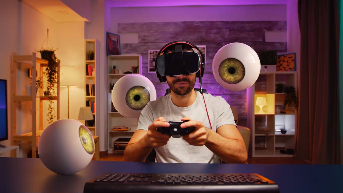 A gamer wearing headphones and a gray shirt intensely plays a video game with large, surreal eyeball decorations around them.