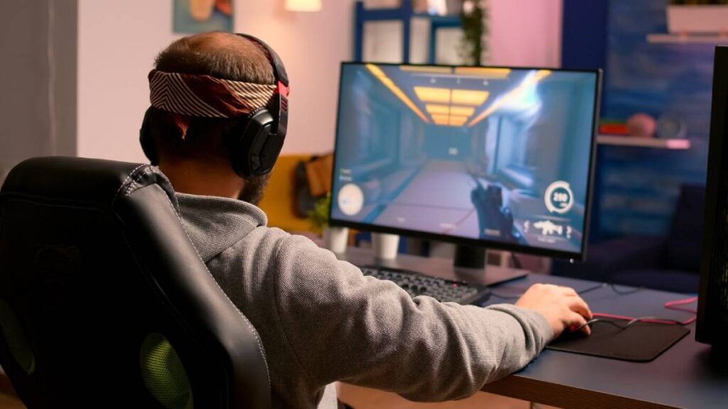 A man wearing headphones plays a video game on a computer, focused on a colorful screen in a cozy gaming setup.