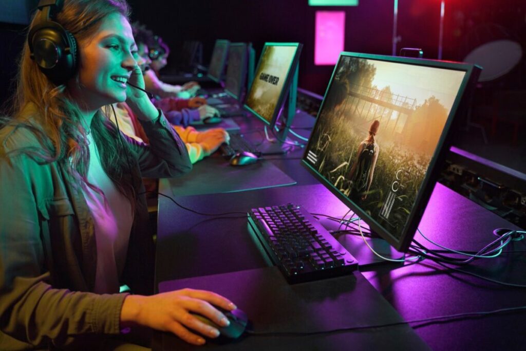 A gamer wearing headphones interacts with a computer, displaying a scenic game environment, amidst colorful, dimly lit surroundings.