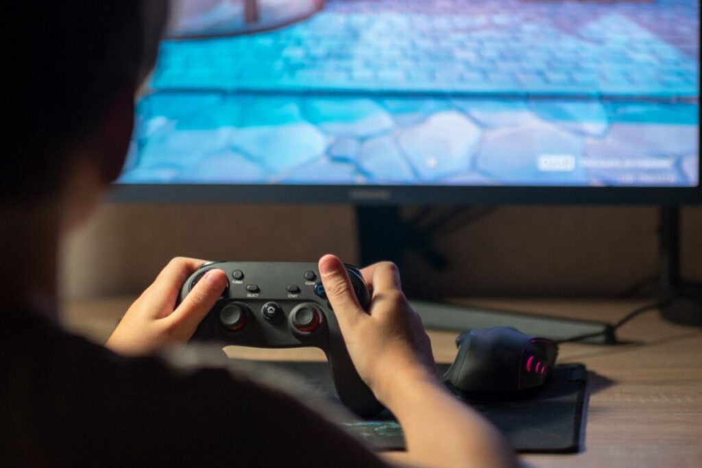 A person holds a game controller in front of a computer screen displaying a colorful video game environment.