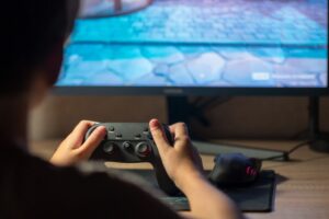 A person holds a game controller in front of a computer screen displaying a colorful video game environment.