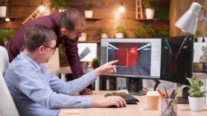 Two men collaborate at a desk, analyzing a 3D design of a brightly lit corridor with red doors on dual monitors.