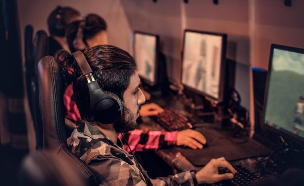 A group of gamers wearing headsets focus intently on their computer screens in a dimly lit gaming lounge.
