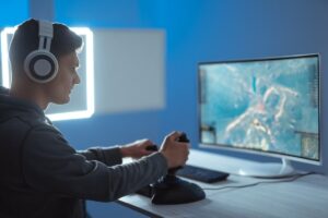 A gamer wearing headphones, focused on a monitor displaying an action-packed game, using a joystick and keyboard in a dimly lit room.