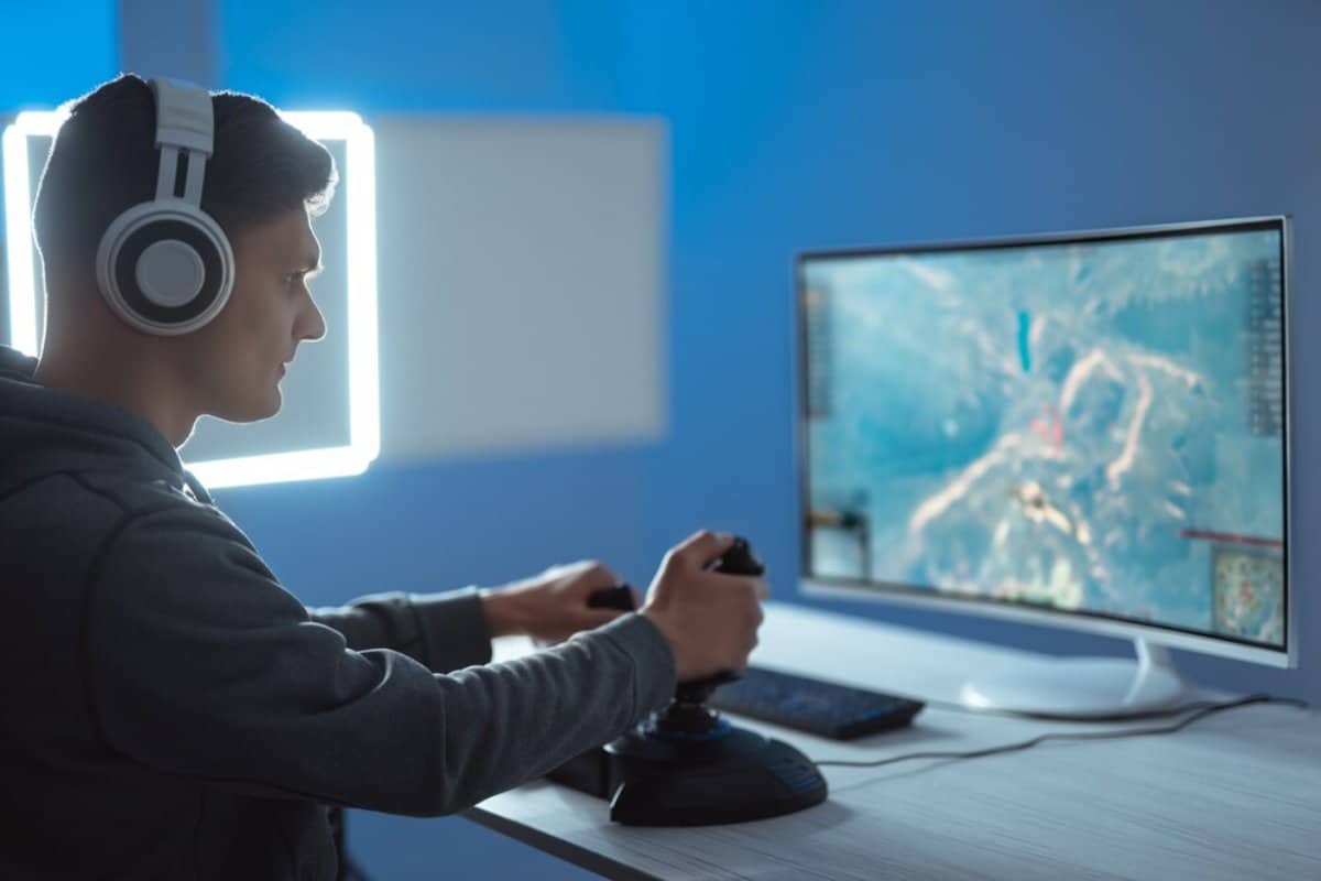 A gamer wearing headphones, focused on a monitor displaying an action-packed game, using a joystick and keyboard in a dimly lit room.