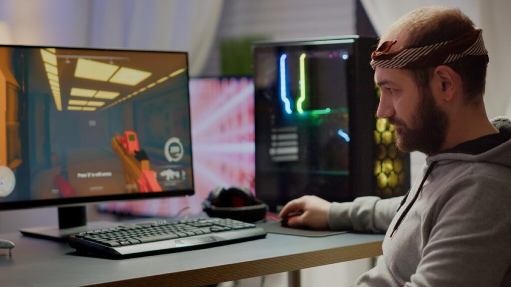A person sitting at a desk gaming on a computer, with a monitor displaying a colorful first-person shooter game.