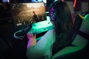 A gamer with long hair wears headphones and plays a video game featuring a character in a scenic outdoor environment.