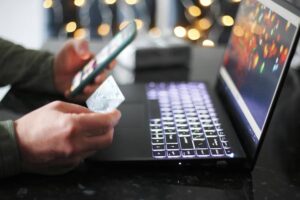 A person holding a smartphone and a credit card, preparing to make an online purchase on a laptop with soft, blurred lights in the background.