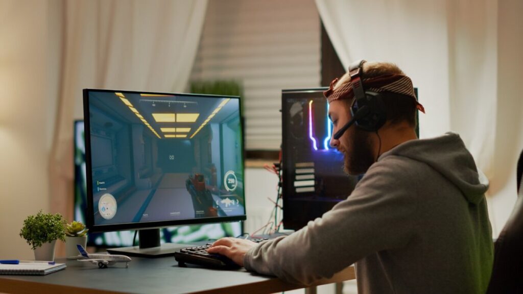 A gamer wearing headphones is intensely playing a first-person shooter game on a large monitor, surrounded by colorful LED-lit equipment.