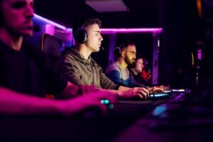 Group of gamers focused on their screens in a dimly lit room with colorful LED lighting, using headsets and gaming keyboards.
