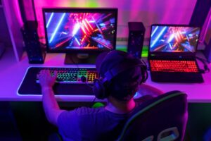 A gamer in a chair, wearing headphones, plays on a desktop and laptop, surrounded by vibrant neon lighting.